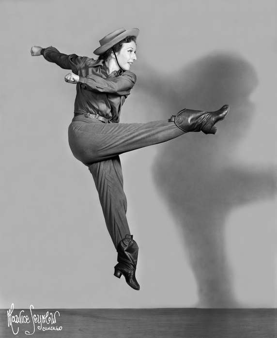 Agnes de Mille in Western inspired garb kicks high in the air. Her hands swing to the side of her.